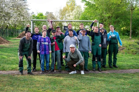 Gruppe von zw&ouml;lf Personen verschiedenen Alters auf einer Wiese vor einem Fu&szlig;balltor. Einige tragen Freizeitkleidung, andere gr&uuml;ne Arbeitskleidung. Drei Personen halten Werkzeuge wie Rechen und Besen. Im Hintergrund bl&uuml;hende B&auml;ume und B&uuml;sche. Klarer Himmel und Fr&uuml;hlingseindruck.
