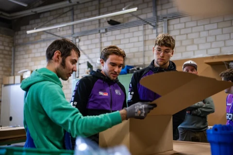 Vier Personen in einer Werkstatt mit gemauerten W&auml;nden und industrieller Beleuchtung; eine Person in gr&uuml;ner Kapuzenjacke faltet einen gro&szlig;en Karton. Zwei Personen tragen lila-schwarze Jacken mit Logos, die vierte Person ist teilweise verdeckt. Auf einem Tisch vor ihnen ist eine blaue Plastikbox sichtbar.