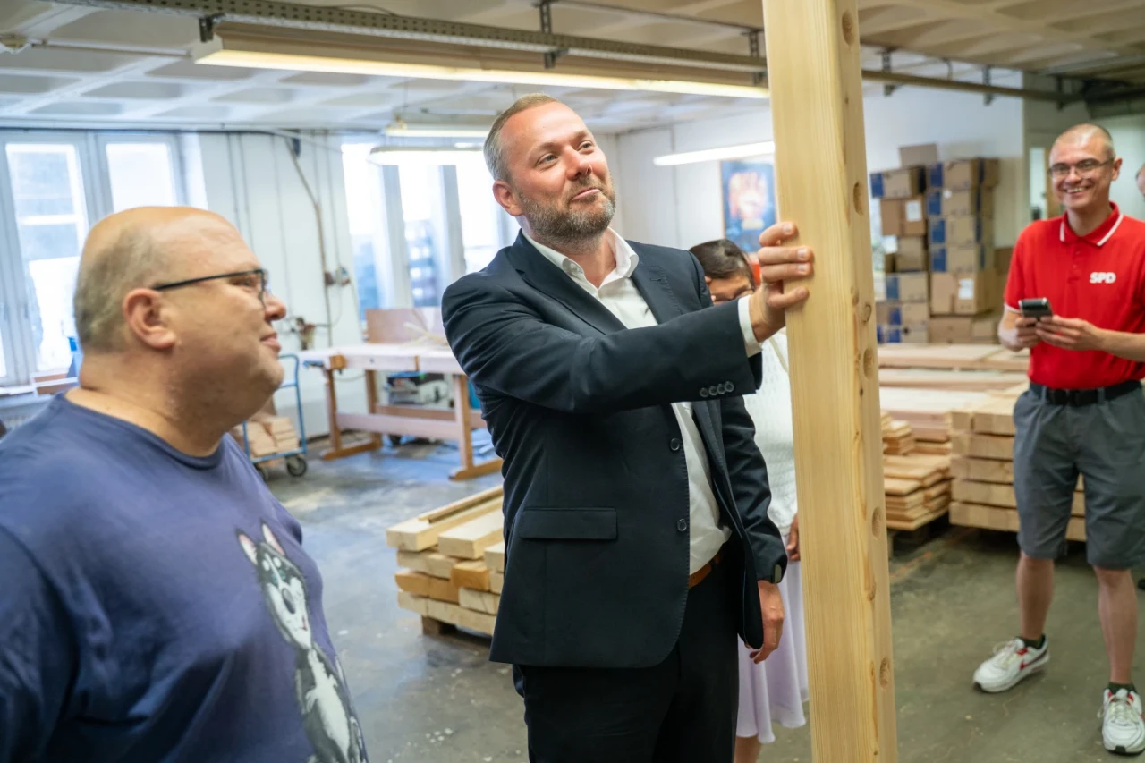 Mann im Anzug h&auml;lt ein vertikal stehendes Holzbrett in einer Werkstatt. Links daneben ein Mann im blauem T-Shirt mit Katzenmotiv. Rechts stehen ein Mann in rotem Polohemd mit SPD-Logo und ein verschwommen sichtbarer Mann. Im Raum sind Fenster, Arbeitsfl&auml;chen und gestapelte Holzlatten sichtbar.