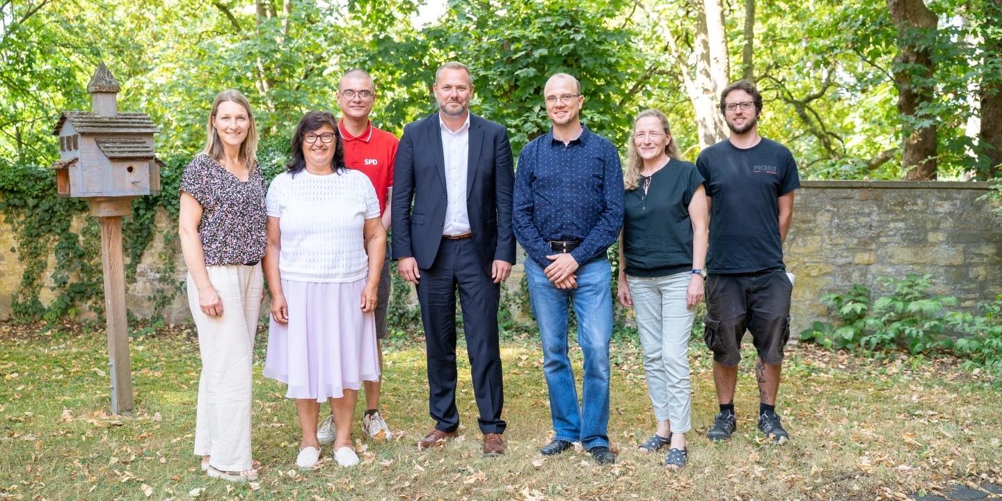 Sieben Personen stehen in einer Reihe auf einer grasbewachsenen Fl&auml;che mit Laub. Im Hintergrund stehen gr&uuml;ne B&auml;ume und eine Steinmauer. Links steht ein brauner h&ouml;lzerner Vogelhausst&auml;nder. Die Personen tragen unterschiedliche Kleidung, darunter Hemd, Bluse, T-Shirt und Anzug. Klare Lichtverh&auml;ltnisse deuten auf sonniges Wetter hin.