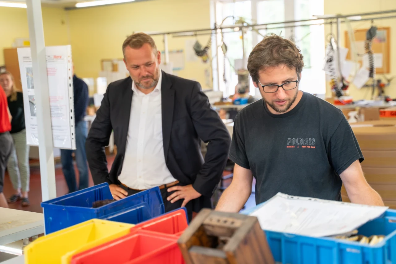 Zwei M&auml;nner in einem B&uuml;ro- oder Werkstattbereich stehen vor mehreren bunten Kunststoffkisten, die auf einem Tisch stehen. Der linke Mann tr&auml;gt einen dunklen Anzug und ein wei&szlig;es Hemd, der rechte ein schwarzes Shirt mit einem dezenten Aufdruck. Der Raum im Hintergrund ist hell beleuchtet, mit gelben W&auml;nden, gro&szlig;en Fenstern und B&uuml;roartikeln an einer Pinnwand.