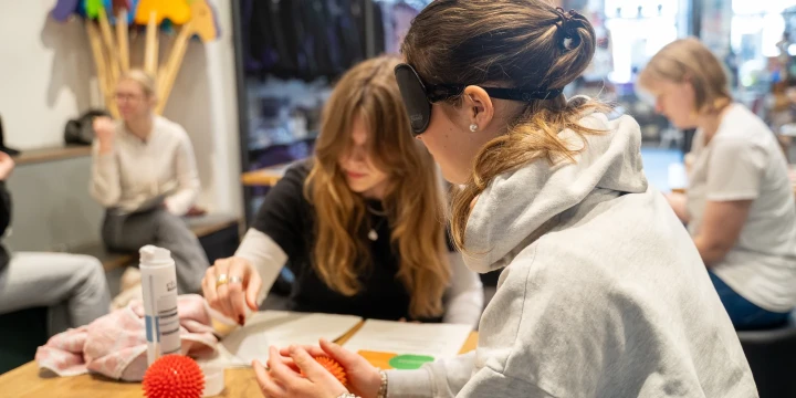 Personen sitzen an einem Holztisch, zwei Frauen im Vordergrund, wobei eine mit wei&szlig;em Hoodie eine rote Noppenkugel h&auml;lt und die andere auf ein Papier deutet. Auf dem Tisch liegen Bastelmaterialien, eine Stoffrolle und Kleber. Im Hintergrund unscharfe Personen, Regale mit bunten Gegenst&auml;nden und Fensterfront mit Tageslicht.
