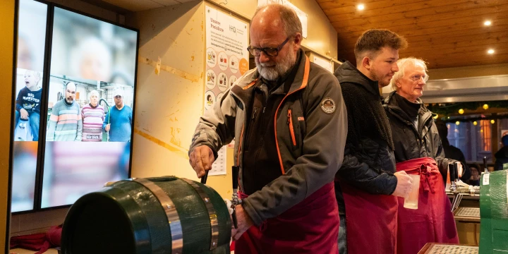 Ein Mann in grauer Jacke und roter Sch&uuml;rze zapft Bier aus einem gr&uuml;n lackierten Holzfass. Hinter ihm stehen zwei weitere M&auml;nner in roten Sch&uuml;rzen. Im Hintergrund eine beleuchtete Holzdecke, Bildschirme mit Fotos und Informationsplakate.