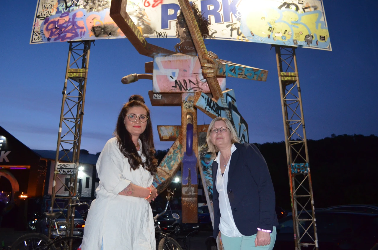 Metallskulptur in Menschengestalt mit ausgestreckten Armen, verziert mit Graffiti, umgeben von zwei Metalltr&auml;gern mit ebenfalls buntem Graffiti. Dar&uuml;ber ein beleuchtetes Schild mit der Aufschrift &bdquo;PARK&ldquo;, ebenfalls bemalt. D&auml;mmerungshimmel im Hintergrund, parkende Autos und beleuchtete Geb&auml;ude sichtbar.