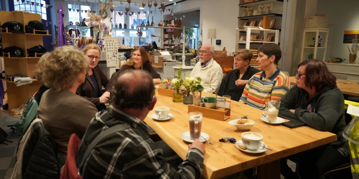 Eine Gruppe von sieben Personen sitzt an einem langen Holztisch in einem hell beleuchteten Raum mit moderner Dekoration. Auf dem Tisch stehen mehrere Kaffeetassen, Gl&auml;ser mit Getr&auml;nken, eine Vase mit Blumen und einige kleine Schalen. Im Hintergrund befinden sich Regale mit dekorativen Objekten, B&uuml;chern und weiteren Produkten, sowie eine Theke.