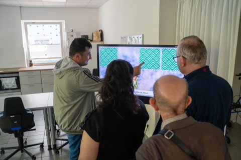 Vier Personen stehen vor einem gro&szlig;en Bildschirm in einem hellen Raum. Eine Person links deutet mit der Hand auf den Bildschirm, der gr&uuml;ne Muster auf blau-wei&szlig;em Hintergrund zeigt. Im Raum befinden sich eine K&uuml;chenzeile links mit Schr&auml;nken und eine schwarze B&uuml;rostuhl-Sitzgruppe.
