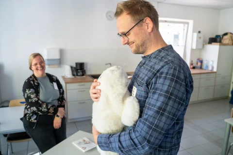 Mann mit Brille, kariertem Hemd und Namensschild h&auml;lt ein wei&szlig;es, pl&uuml;schiges Roboter-Tier, &auml;hnlich einem Seehund, in einem B&uuml;ro mit K&uuml;chenzeile. Im Hintergrund sitzt eine l&auml;chelnde Frau mit Brille und Bluse sowie einer Blumenjacke an einem Tisch. Auf dem Tisch ein offen liegendes Buch.