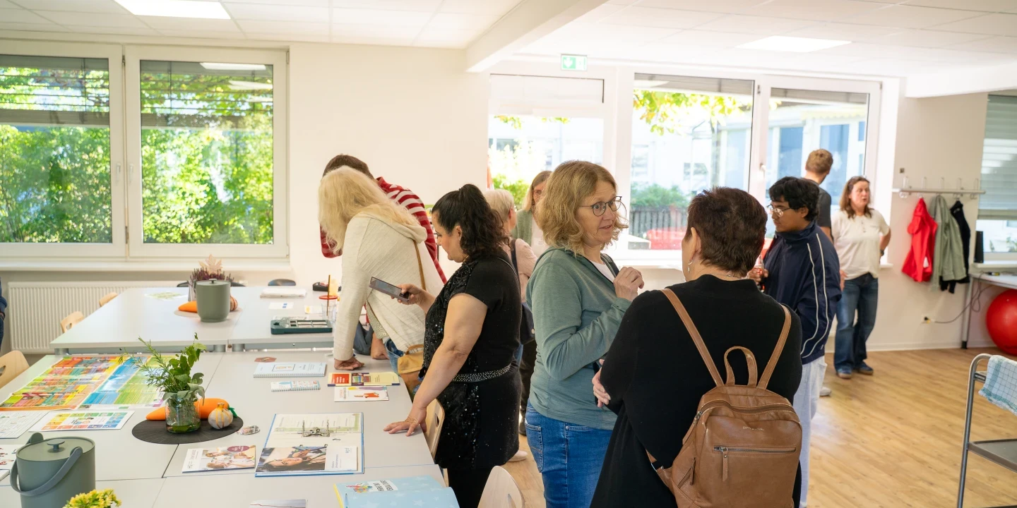 Personen in einem hellen Raum mit wei&szlig;en W&auml;nden und gro&szlig;en Fenstern, die durch Tageslicht beleuchtet werden. Auf mehreren Tischen liegen Zeitschriften, B&uuml;cher, Flyer und Dekorationsartikel wie Pflanzen und Fr&uuml;chte. Einige Personen betrachten die Auslage, andere unterhalten sich. Im Hintergrund eine Garderobe und ein rollbarer Wagen.
