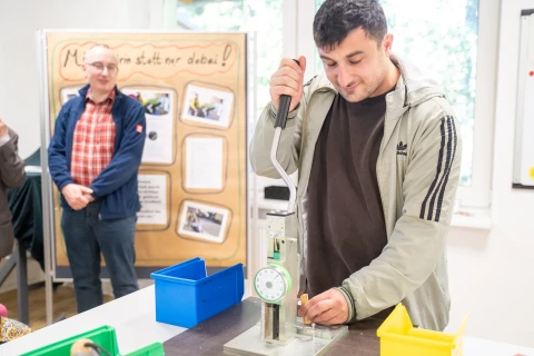 Mann mit beigem Hoodie bedient eine mechanische Presse auf einem wei&szlig;en Tisch mit gelbem, gr&uuml;nem und blauem Beh&auml;lter. Im Hintergrund ein Mann mit kariertem Hemd und Poster mit Text und Fotos, helle Fenster rechts.