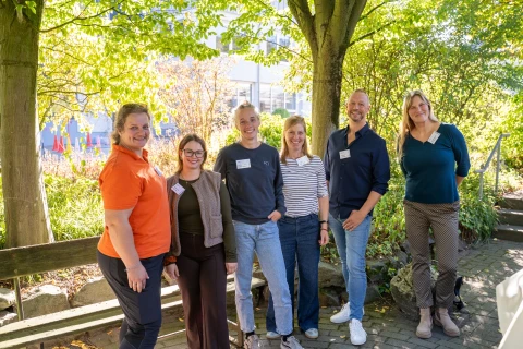 Sechs Personen stehen nebeneinander in einer gr&uuml;nen Gartenumgebung mit B&auml;umen und B&auml;umen, teils aufrecht, teils l&auml;chelnd. Im Hintergrund sind bunte Pflanzen, ein Geb&auml;udeteil mit Glasfassade und rote Stuhlformen erkennbar. Lichtstrahlen durchbrechen die Baumkronen.