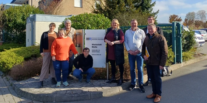 Sechs Personen stehen auf einem Gehweg vor einem Schild mit der Aufschrift "Werkstatt Melle". Das Schild ist wei&szlig; mit blauer Schrift und steht rechts vor einer gr&uuml;nen Hecke und einem Zaun. Dahinter ein Backsteinhaus mit schr&auml;gem Dach und B&auml;umen. Am rechten Bildrand parken Autos unter blauem Himmel.