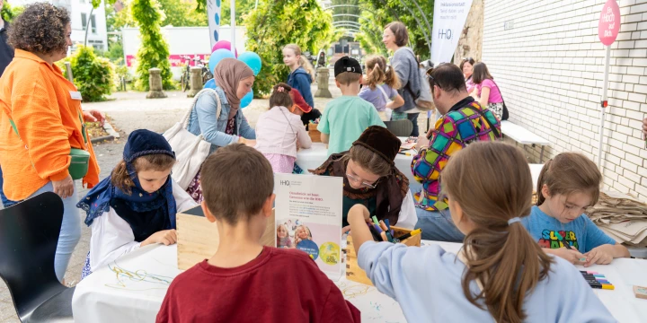 Kinder und Erwachsene sitzen und stehen an einem gro&szlig;en wei&szlig;en Tisch unter einem Pavillon mit wei&szlig;em Dach, nutzen Bastelmaterialien und besch&auml;ftigen sich kreativ. Links bunte Luftballons und eine Frau in orangener Jacke, hinten B&auml;ume und ein Geb&auml;ude vor einer wei&szlig;en Wand.