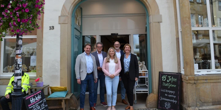 F&uuml;nf Personen stehen vor einem Geb&auml;ude mit offenem Eingang und dem Schriftzug "Stadtgalerie" &uuml;ber dem T&uuml;rbogen. Links ein Blumentopf mit pinken Blumen an einer dekorierten Laterne, rechts eine Tafel mit der Aufschrift "15 Jahre StadtGalerie, Jubil&auml;umsausstellung". Hausnummer 13 auf der Fassade.