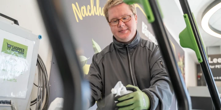 Person in grauer Arbeitsjacke mit gr&uuml;nem Handschuh wischt die Griffstange eines Einkaufswagens ab. Im Hintergrund eine Werbetafel mit gelbem Text: "f&uuml;r mehr Nachhaltigkeit". Links ein wei&szlig;er Beh&auml;lter mit Aufschrift: "SHOPPER Reinigung Carts". Teile eines Ladeneingangs rechts erkennbar.