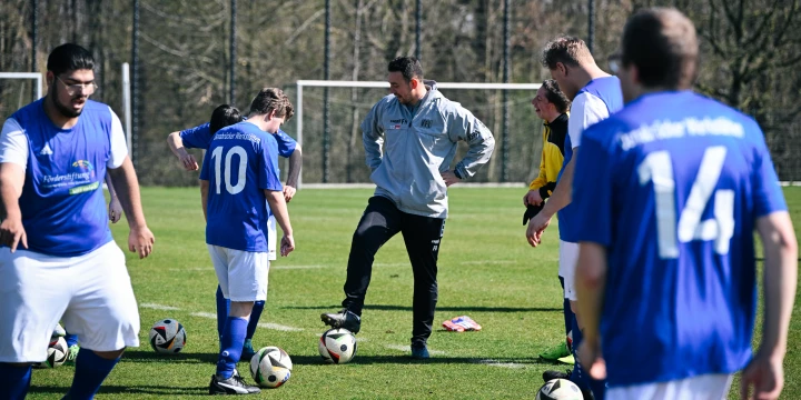 Gruppe von sechs Personen auf einem Fu&szlig;ballfeld; f&uuml;nf Spieler in blauen Trikots und wei&szlig;en Hosen, eine Person im grauen Trainingsanzug mit Ball unter dem Fu&szlig;. Auf dem Rasen verteilt mehrere Fu&szlig;b&auml;lle, im Hintergrund Tor mit Netz und B&auml;ume.