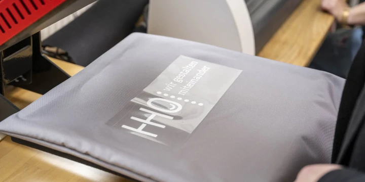 Graues Textil auf einer rechteckigen Heizpresse mit transparentem Druckmotiv, das die Schrift "HHO, wir gestalten miteinander" und wei&szlig;e grafische Elemente zeigt. Im Hintergrund sind Holztisch, rote und silberne Maschinenteile sowie eine teilweise sichtbare Hand erkennbar.