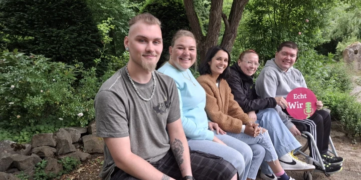 F&uuml;nf Personen sitzen nebeneinander auf einer Holzbank in einem Park. Im Hintergrund B&auml;ume und Str&auml;ucher, im Vordergrund ein unbefestigter Boden. Eine Person h&auml;lt ein pinkfarbenes Schild mit der Aufschrift "Echt Wir".