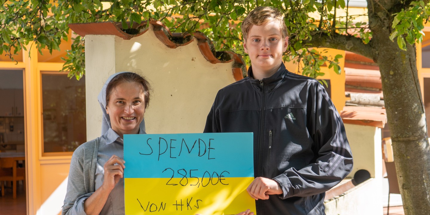 Zwei Menschen halten ein blau-gelbes Schild mit der Spendensumme hoch.