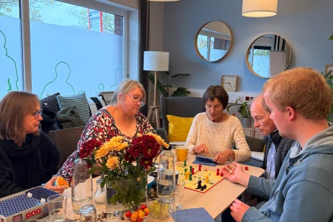 F&uuml;nf Personen unterschiedlichen Alters sitzen an einem rechteckigen Holztisch und spielen ein Brettspiel. Auf dem Tisch stehen eine Vase mit farbigen Blumen, Gl&auml;ser, Wasserflaschen, ein Teller mit Geb&auml;ck und Obst sowie eine orangefarbene Tasse. Im Hintergrund Spiegel, Pflanzen und H&auml;ngelampen; durch Fenster rechts sichtbare Au&szlig;enwelt.