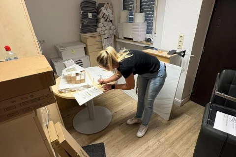 Frau mit blonden Haaren, schwarzem T-Shirt und blauer Jeans beugt sich &uuml;ber einen runden wei&szlig;en Tisch und schreibt auf Papieren. Auf dem Tisch mehrere Kartons, Dokumente und B&uuml;robedarf. Im Raum stapeln sich Kartons und Akten, links ein Regal, rechts ein schwarzer B&uuml;rostuhl, helle Holzb&ouml;den und wei&szlig; gestrichene W&auml;nde.