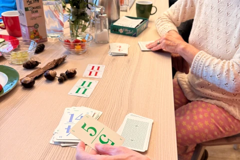 Personen unterschiedlichen Alters sitzen an einem langen Holztisch und spielen Karten. Drei Karten mit Zahlen 14 und 11 in Rot und Gr&uuml;n liegen sichtbar aus. Auf dem Tisch stehen eine Thermoskanne, diverse Tassen, Blumenvasen mit bunten Blumen, eine Schale mit N&uuml;ssen und Gl&auml;ser mit kleinen Snacks. Im Hintergrund sind weitere Personen und Dekorationen zu sehen.