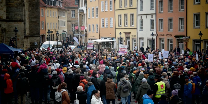 Menschenmenge auf einem gepflasterten Platz umgeben von mehrst&ouml;ckigen H&auml;usern in Pastellfarben; viele tragen Winterkleidung. Einige Personen halten Plakate mit unleserlichem Text. Rechts sind gelb gekleidete Ordner sichtbar. Links ein Kirchengeb&auml;ude und ein blauer Pavillon.