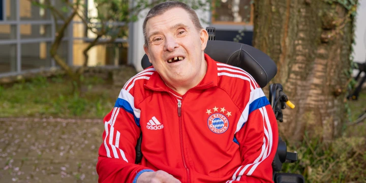 Person im Rollstuhl mit roter Adidas-Trainingsjacke des FC Bayern M&uuml;nchen, auf der linke Brustseite das Vereinslogo mit vier Sternen. Im Hintergrund ein Baum, Geb&auml;udeteil mit Fenstern und gr&uuml;ne Pflanzen. Boden mit vereinzelten rosa Bl&uuml;tenbl&auml;ttern.