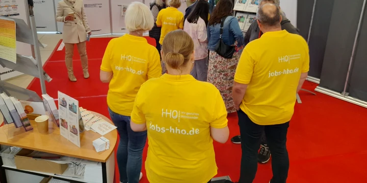 Personengruppe in gelben T-Shirts mit der Aufschrift "jobs-hho.de" auf einem roten Boden, in einer Messehalle. Im Hintergrund stehen weitere Personen vor Info-St&auml;nden mit Plakaten, Flyern und Trennw&auml;nden. Links Informationsmaterial, rechts ein Aufsteller mit dem Logo "HHO".