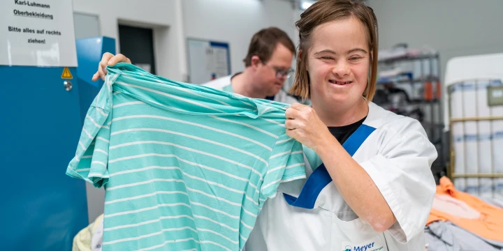 Person in wei&szlig;er Arbeitsbekleidung mit blauem Firmenlogo "Meyer" h&auml;lt ein t&uuml;rkisfarbenes gestreiftes T-Shirt mit beiden H&auml;nden vor sich. Im Hintergrund blauer Schrank mit Schild, W&auml;schek&ouml;rbe und weitere Kleidungsst&uuml;cke.