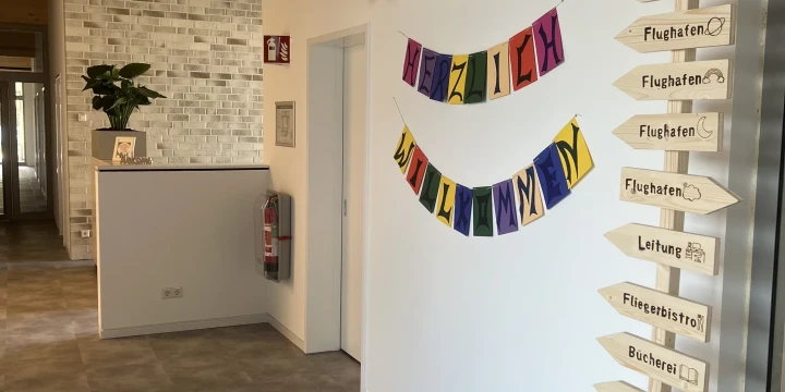Blick in einen Flur mit grauem Boden und wei&szlig; gestrichenen W&auml;nden. An der rechten Wand bunte Deko mit Wolken, Sonne, Regenbogen und zwei Girlanden mit dem Text "Herzlich Willkommen". Daneben ein Wegweiser aus Holz in einem Topf mit Steinen, dessen Schilder Begriffe wie "Flughafen", "Leitung", "Caf&eacute;" und "WC" zeigen. Links ein niedriger Tresen mit einer Pflanze und ein Feuerl&ouml;scher an der Wand.