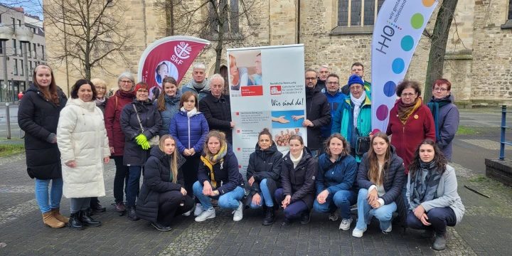 Gruppe von 22 Personen, &uuml;berwiegend in Winterkleidung, vor einem historischen Geb&auml;ude mit gro&szlig;en gotischen Fenstern. Im Hintergrund zwei Roll-Up-Banner mit Logos und Texten sowie zwei bedruckte Flaggen. Pflastersteine bedecken den Boden, kahle B&auml;ume sind sichtbar.