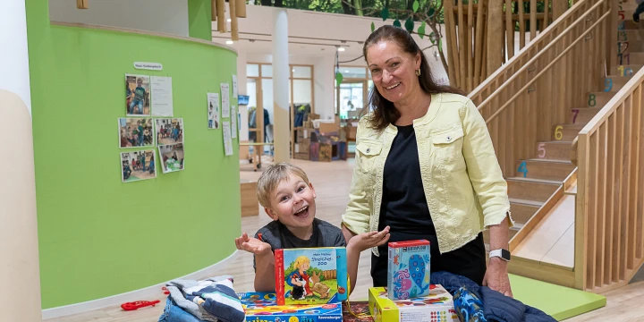 Ein Kind und eine Frau stehen hinter einem Holztisch, der mit Gesellschaftsspielen, Malutensilien und gefalteten Stoffteilen wie Kleidung belegt ist. Im Hintergrund ist ein Raum mit Holzelementen, einer Treppe mit bunten Zahlen und gr&uuml;nen W&auml;nden zu sehen, dekoriert mit Fotos und Zeichnungen.