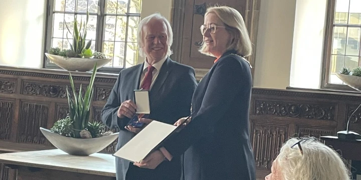 Ein Mann in Anzug mit roter Krawatte h&auml;lt eine Medaille, daneben steht eine Frau in dunklem Hosenanzug mit ge&ouml;ffnetem Ordner. Im Raum Holzverkleidung, gro&szlig;e Fenster mit bleiverglastem Glas, Topfpflanzen auf einem Holztisch, Gem&auml;lde eines Mannes &uuml;ber einer T&uuml;r. Im Vordergrund mehrere sitzende Personen.