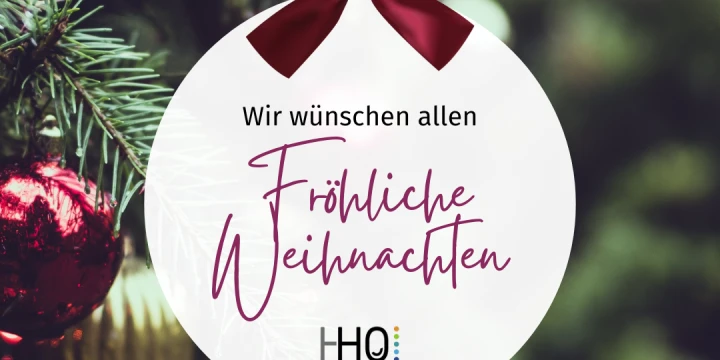 Runder, wei&szlig;er Christbaumanh&auml;nger mit roter Schleife, beschriftet mit "Wir w&uuml;nschen allen Fr&ouml;hliche Weihnachten" in schwarzer und roter Schrift. Darunter das Logo "HHO" mit buntem Punktmuster. Im Hintergrund unscharfe gr&uuml;ne Tannenzweige mit roten und goldenen Kugeln.