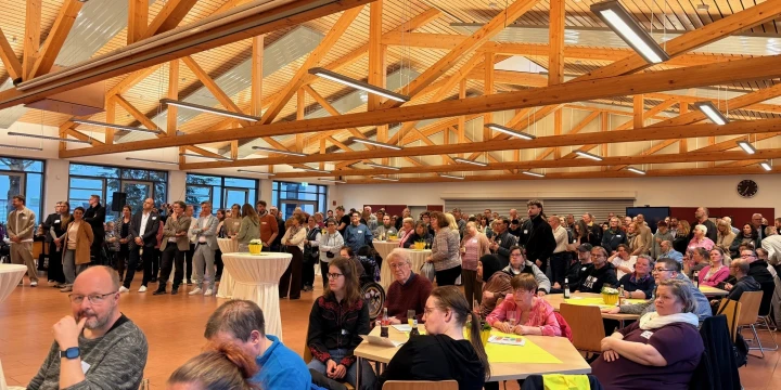 Gro&szlig;er Saal mit Holzdeckenbalken, gef&uuml;llt mit vielen Menschen. Personen sitzen an Tischen im Vordergrund, einige mit Getr&auml;nken und Papieren. Im Hintergrund stehen gr&ouml;&szlig;ere Menschengruppen, daneben Stehtische. Tageslicht f&auml;llt durch gro&szlig;e Fenster auf der linken Seite ein, eine Wanduhr h&auml;ngt rechts hinten.