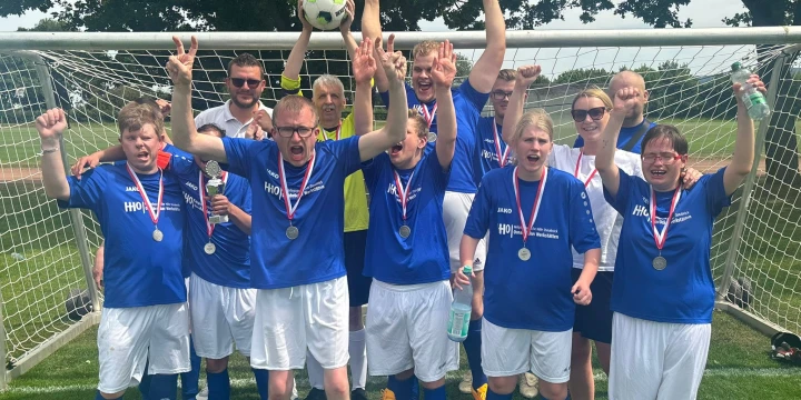 Eine Gruppe von elf Fu&szlig;ballspielern in blauen Trikots und wei&szlig;en Shorts steht vor einem Fu&szlig;balltor auf einem Rasenplatz. Alle tragen Medaillen um den Hals, einige halten Pokale, eine Person h&auml;lt einen Fu&szlig;ball &uuml;ber dem Kopf. Im Hintergrund sind gr&uuml;ne B&auml;ume und ein Feld sichtbar.
