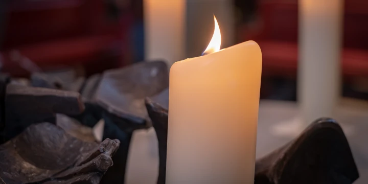 Brennende cremefarbene Kerze mit leichter Flamme, umgeben von dunkelgrauen, geschwungenen Metallelementen. Im Hintergrund unscharf zwei weitere brennende Kerzen und rote Sitzb&auml;nke.
