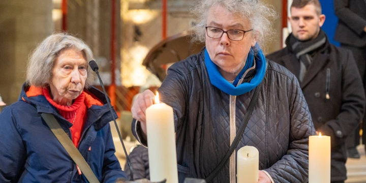 Person in dunkelblauer Steppjacke mit blauer Schalkapuze z&uuml;ndet eine wei&szlig;e, lange Kerze an. Weitere Kerzen und Mikrofon auf Tisch. Im Hintergrund unscharfe, teils verschwommene Personen und kirchliche Innenraumdetails.