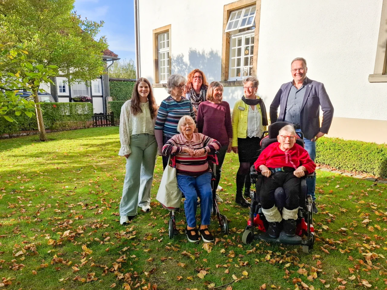 Gruppe von acht Personen im Garten, f&uuml;nf stehend und drei sitzend; zwei Personen in Rollst&uuml;hlen. Vier Frauen, ein Mann, alle l&auml;chelnd. Hintergrund: wei&szlig;es Geb&auml;ude mit gro&szlig;en Fenstern, B&auml;ume und Rasen mit gelbem Herbstlaub.