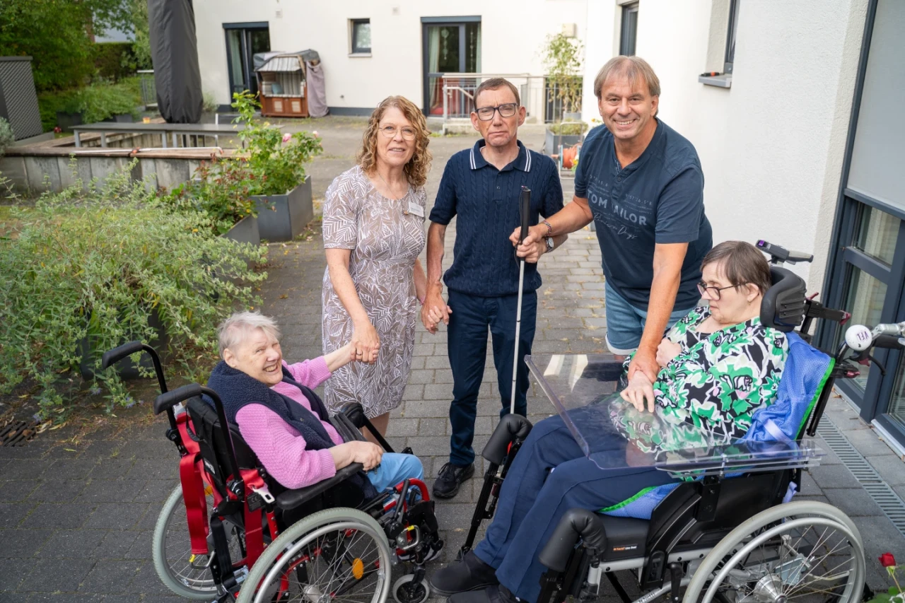 Drei Erwachsene und zwei &auml;ltere Menschen in Rollst&uuml;hlen auf einer gepflasterten Terrasse. Zwei Erwachsene stehen mittig, eine Frau h&auml;lt die Hand einer Rollstuhlfahrerin, ein Mann mit Blindenstock steht daneben, rechts ein Mann in Blau. Im Hintergrund Pflanzen, Geb&auml;ude und ein Whirlpool.