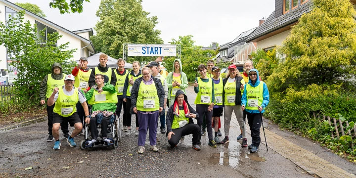 Gruppe von 16 Personen unterschiedlicher Altersgruppen in gelben Warnwesten mit "HHO Runners"-Aufdruck, stehend und sitzend vor einem Startbanner. Die meisten Personen tragen Sportkleidung und l&auml;cheln, im Hintergrund Wohnh&auml;user, B&uuml;sche und B&auml;ume. Der Boden ist nass.