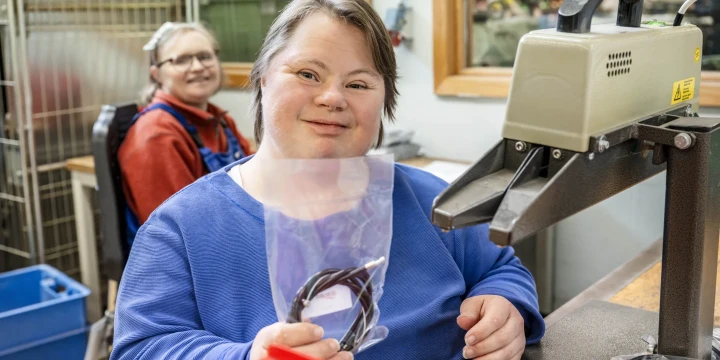 Person in blauer Kleidung h&auml;lt eine transparente Plastikt&uuml;te mit schwarzen Kabeln vor sich. Rechts von der Person ein Schwei&szlig;ger&auml;t mit abgewinkeltem Arm. Im Hintergrund eine weitere Person in Arbeitsbekleidung, Gitterregal und Kisten. Wand mit Fenster rechts.