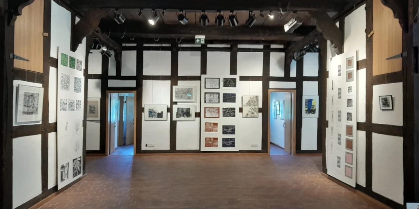 Ausstellungsraum mit wei&szlig;en W&auml;nden und dunklen Holzst&uuml;tzen, dekoriert mit gerahmten Kunstwerken in verschiedenen Gr&ouml;&szlig;en. Boden aus braunen Kacheln, Decke aus dunklem Holz mit Strahlern. T&uuml;ren f&uuml;hren in angrenzende R&auml;ume.