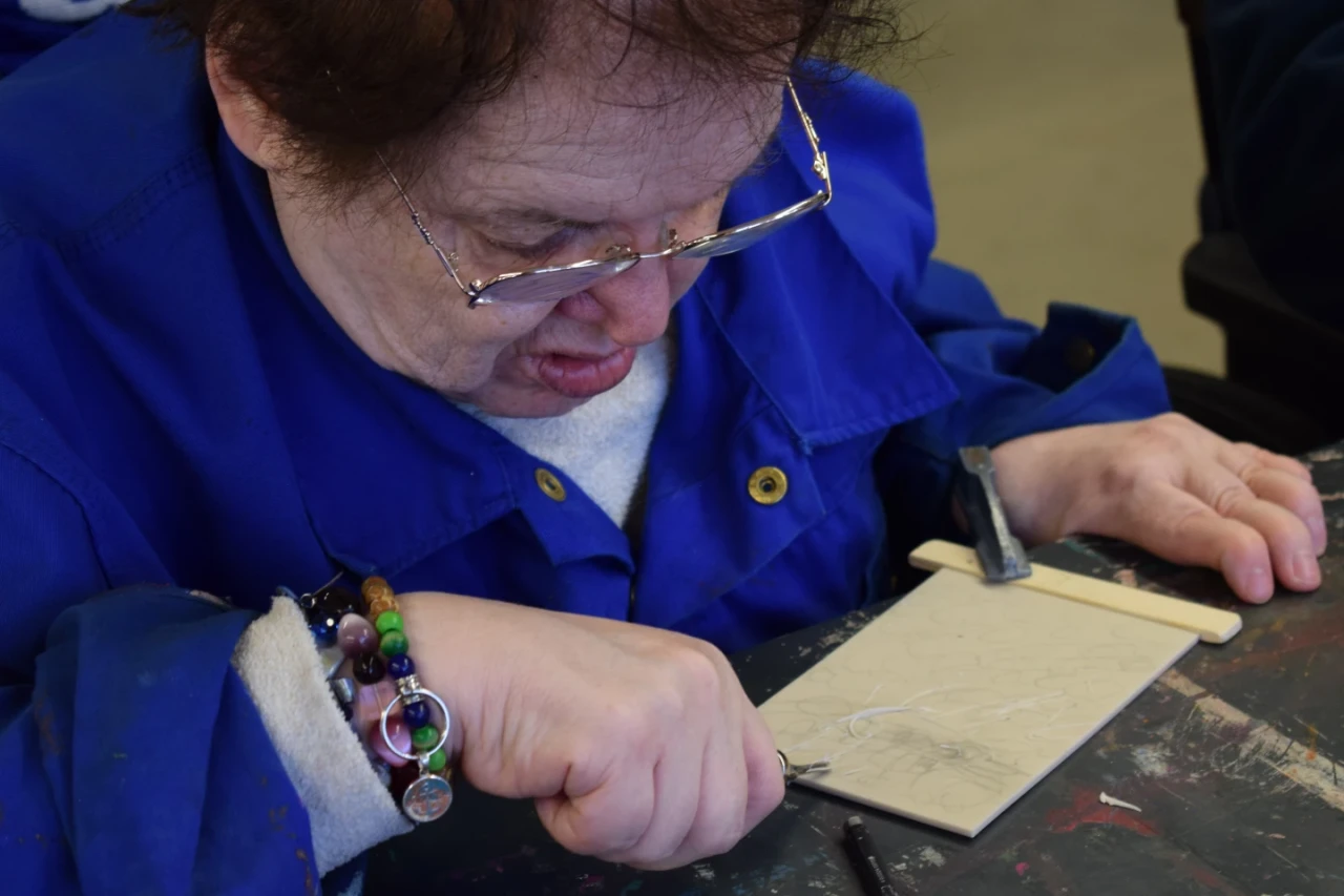 Eine Person mit braunem Haar und Brille graviert ein kleines Muster auf eine rechteckige Oberfl&auml;che aus hellem Material. Sie tr&auml;gt eine blaue Arbeitsjacke und ein buntes Armband. Ein schwarzer Stift und ein Gravierwerkzeug liegen auf einer dunklen Tischplatte.