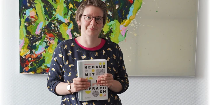 Nora H&uuml;semann schaut in die Kamera und h&auml;lt ein Buch hoch.