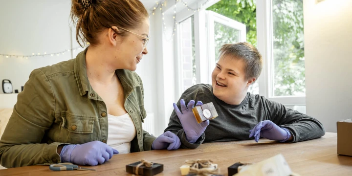Frau in olivgr&uuml;ner Jacke und wei&szlig;em Shirt sowie Mann in grauem Pullover, beide mit lilafarbenen Einmalhandschuhen, sitzen an einem Holztisch. Auf dem Tisch liegen handgefertigte Seifen, ein Ma&szlig;band, eine Schere, ein Notizbuch und ein in Geschenkpapier eingepacktes Paket. Im Hintergrund sind eine wei&szlig;e Wand, eine Lichterkette und ein gro&szlig;es Fenster mit Blick auf gr&uuml;ne B&auml;ume sichtbar.