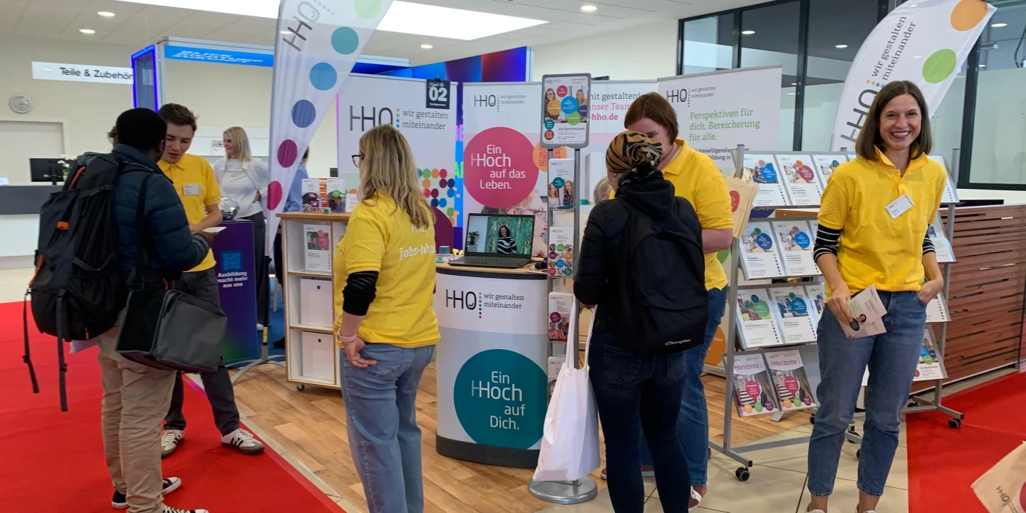 Messestand mit gelbem Bodenrahmen und rotem Teppich, betreut von Personen in gelben T-Shirts. Der Stand zeigt mehrere Brosch&uuml;renst&auml;nder, zwei bedruckte Fahnen und Schilder mit Texten wie "Ein Hoch auf Dich." und "Ein Hoch auf das Leben." Im Hintergrund moderner, heller Raum mit Glaswand und Infotheke.