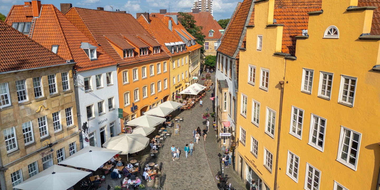 Pflasterstra&szlig;e mit gelben, wei&szlig;en und orangefarbenen Geb&auml;uden mit roten Ziegeld&auml;chern auf beiden Seiten, Stra&szlig;enrestaurants mit wei&szlig;en Sonnenschirmen, darunter ein Brunnen. Menschen spazieren, sitzen und essen. Im Hintergrund modernes Hochhaus und gr&uuml;ne B&auml;ume unter blauem Himmel mit wei&szlig;en Wolken.