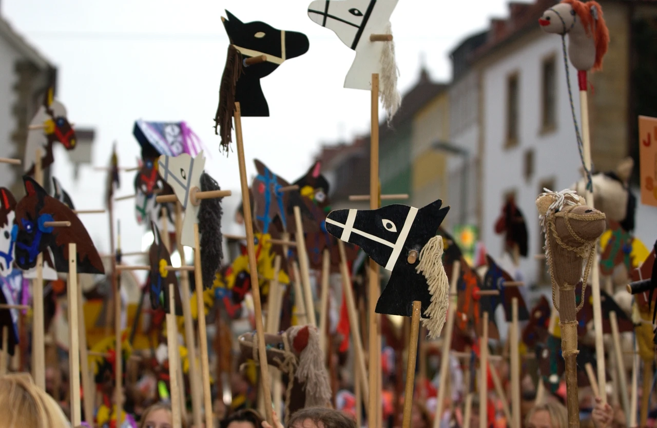 Viele Steckenpferde aus Holz, Stoff und Wolle unterschiedlicher Farben werden von Menschen in einer gro&szlig;en Menschenmenge auf einem &ouml;ffentlichen Platz emporgehalten. Im Hintergrund sind mehrere Geb&auml;ude und unscharfe Details zu sehen.