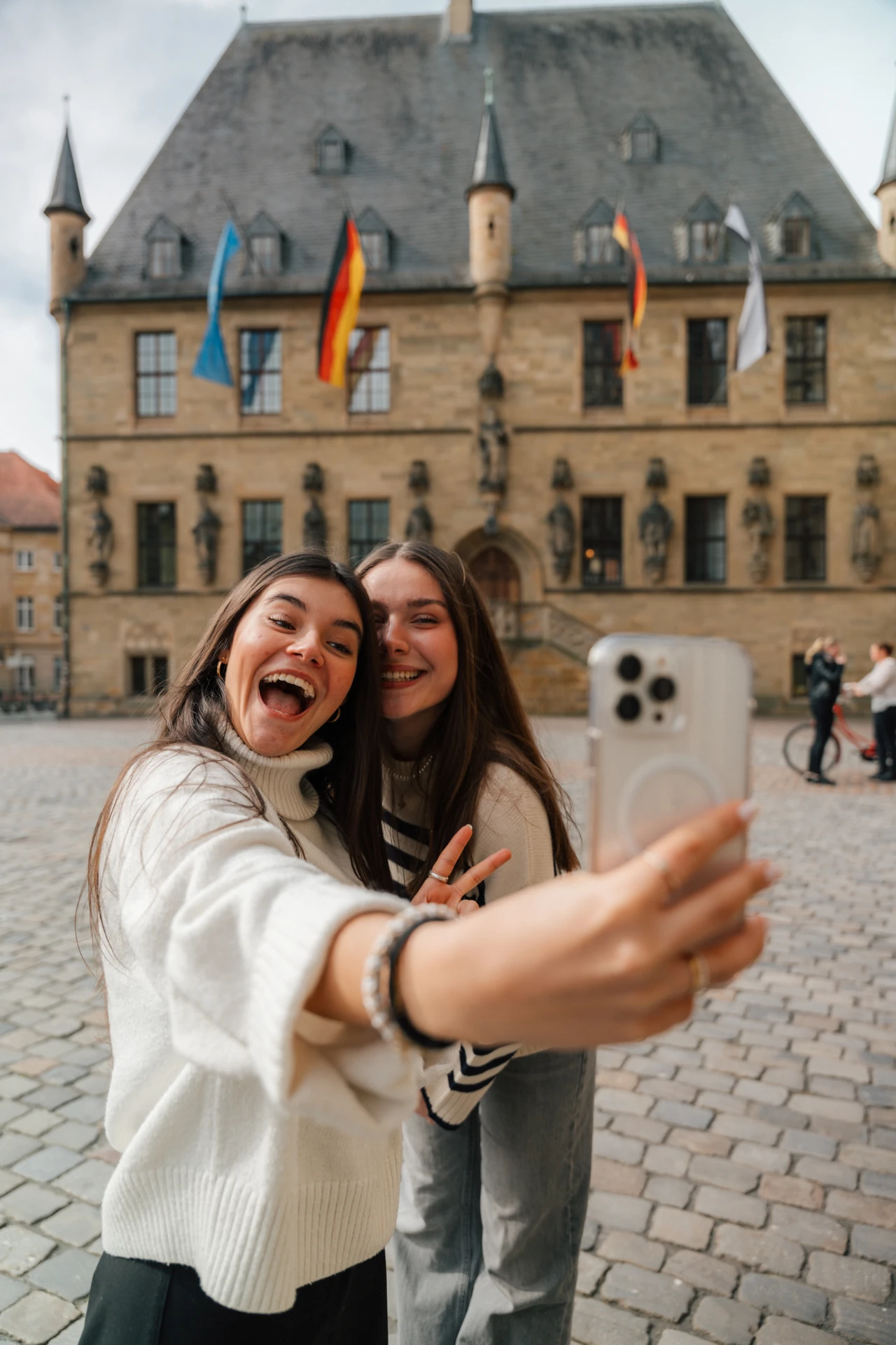 Zwei Personen in heller Kleidung machen ein Selfie mit einem silbernen Smartphone. Im Hintergrund ein historisches Geb&auml;ude aus beigefarbenem Stein mit mehreren Fenstern, T&uuml;rmchen und wehendem Flaggen, darunter deutsche Flaggen und blaue Fahnen. Kopfsteinpflaster bedeckt den Boden, rechts im Bild stehen weitere Menschen.